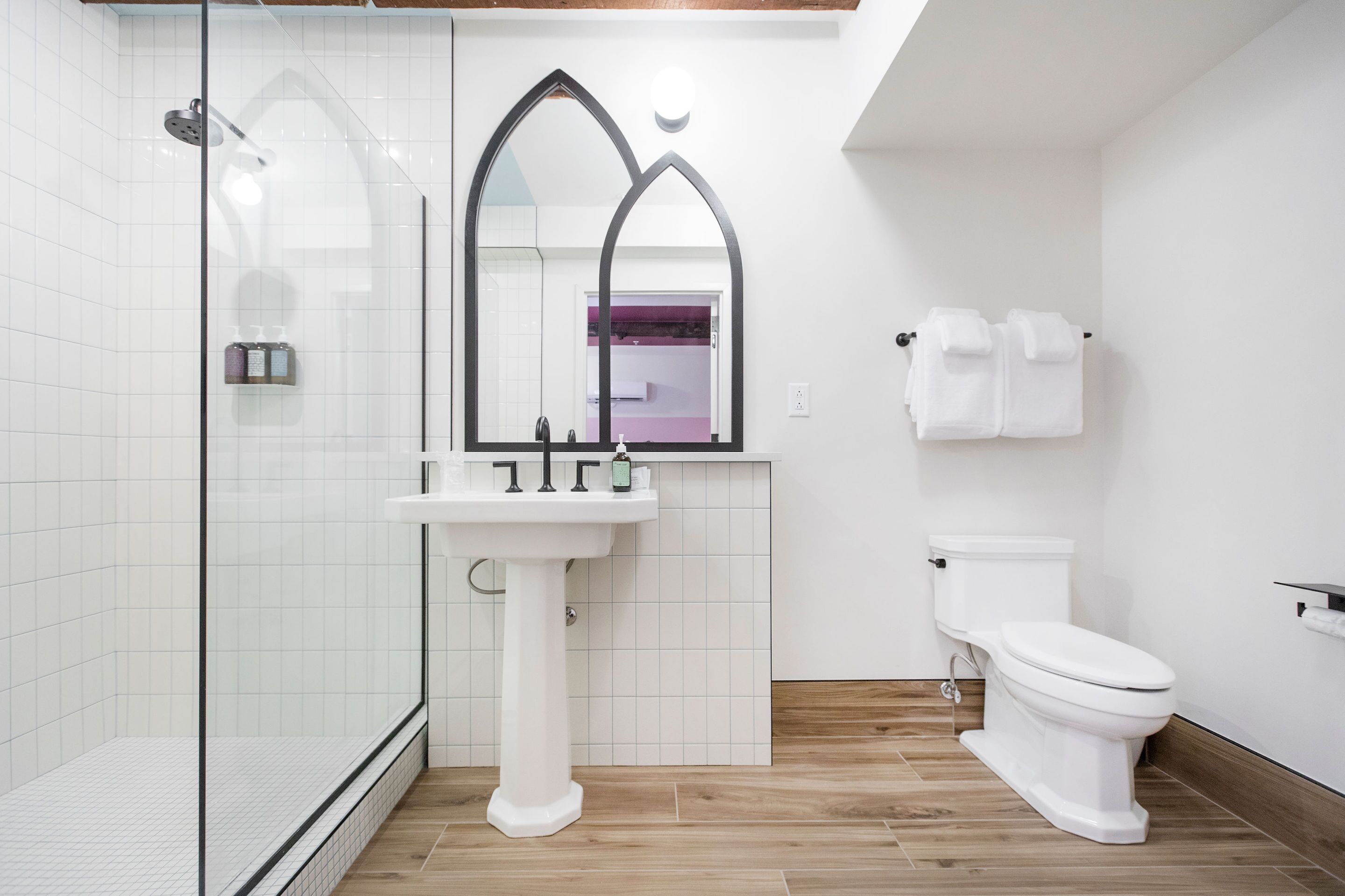 A bathroom with a glass shower enclosure, a pedestal sink, a toilet, and a mirror with a gothic arch design.