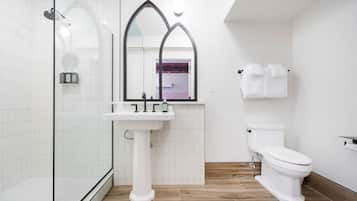 A bathroom with a glass shower enclosure, a pedestal sink, a toilet, and a mirror with a gothic arch design.