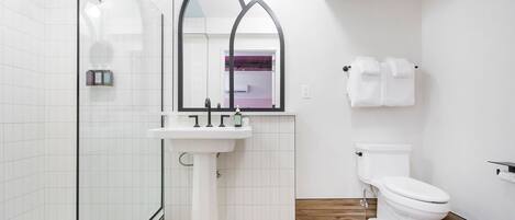 A bathroom with a glass shower enclosure, a pedestal sink, a toilet, and a mirror with a gothic arch design.