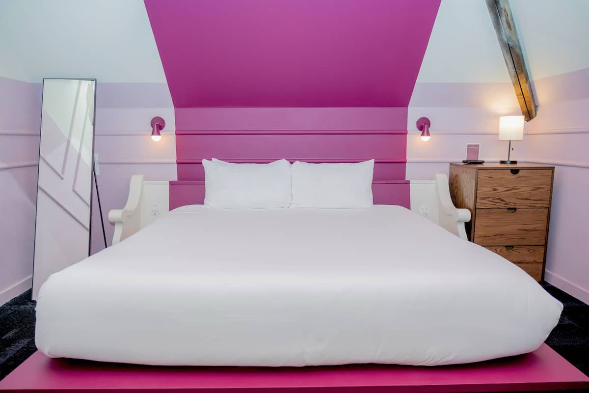Ein großes Bett mit weißen Leinen, eine Holzkommode und zwei Nachttischlampen in einem Zimmer mit rosa und weißen Wänden.