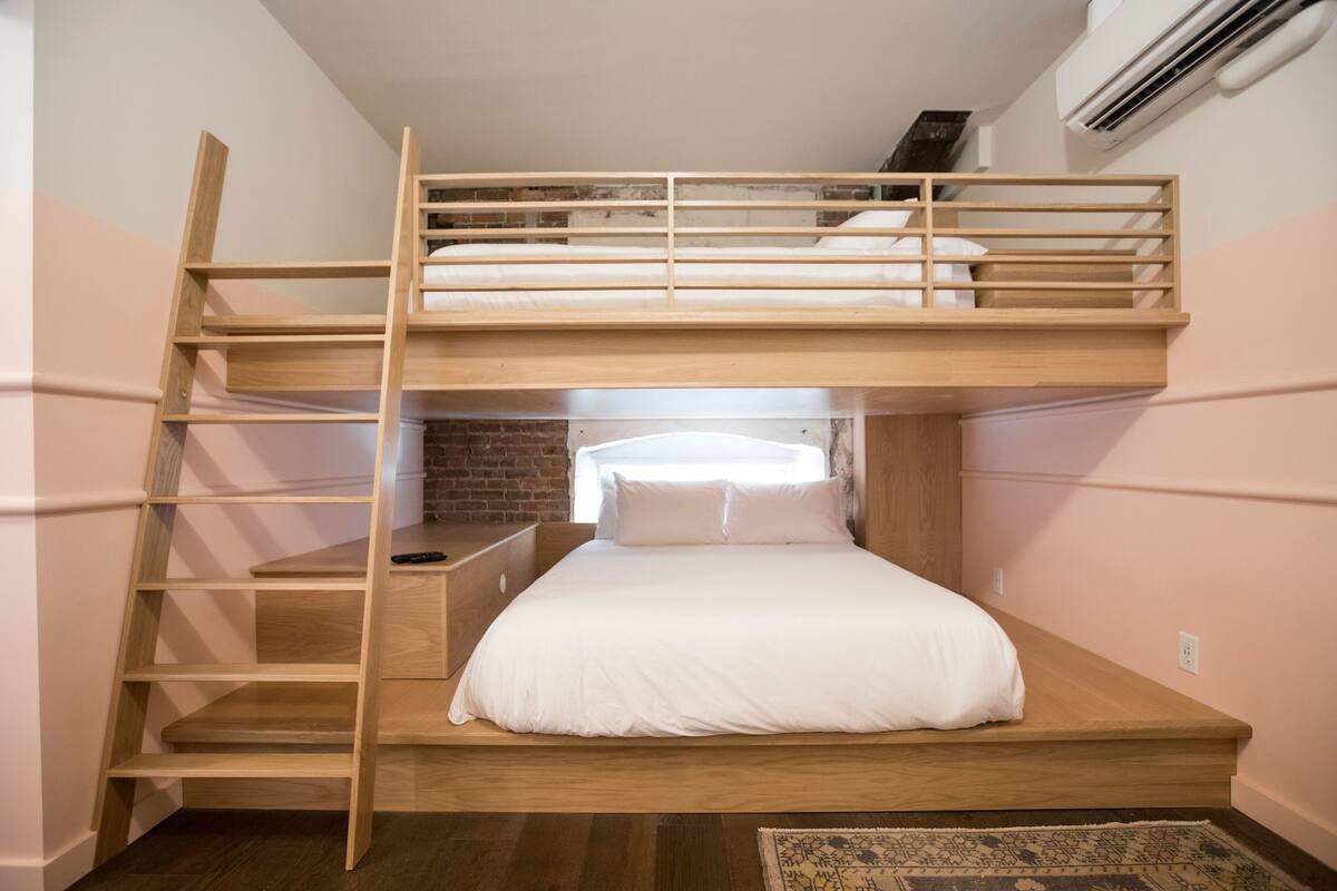 Ein Stockbett mit Holzlade, ein kleiner Schreibtisch und eine Ziegelwand im Hintergrund.