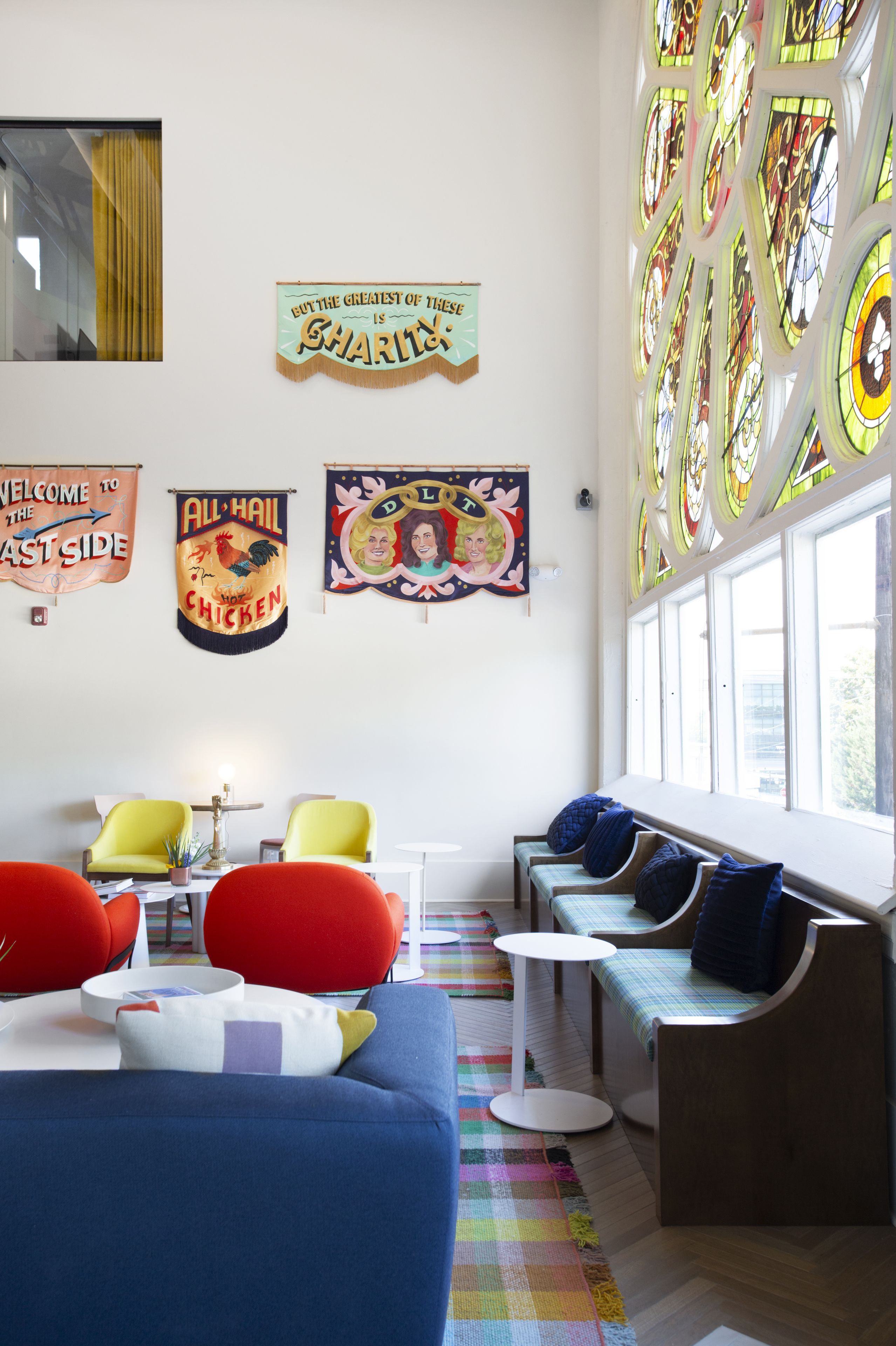 Una habitación con un sofá azul, sillas rojas y un banco con cojines azules. Hay banderines de colores en la pared y un ventanal con vidrieras.