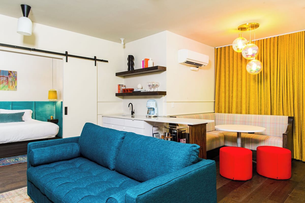 Ein modernes Hotelzimmer mit einem türkisfarbenen Sofa, einer kleinen Küchenzeile, einem Esstisch mit roten Hocker und einem gelben Vorhang im Hintergrund.