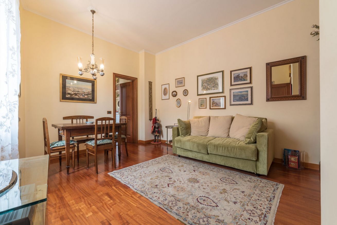 A living room with a green sofa, wooden dining table, chairs, a chandelier, and a patterned rug.