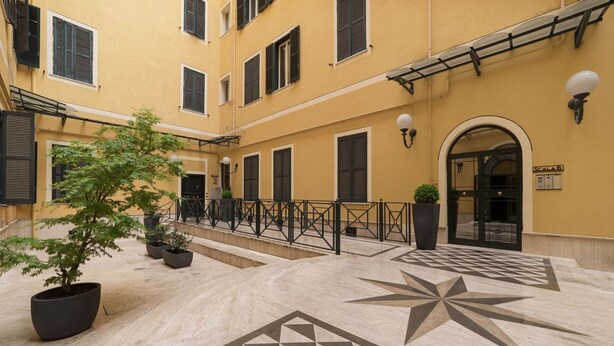 Un patio con un diseño geométrico de azulejos, plantas en macetas y un edificio con varias ventanas y una puerta.
