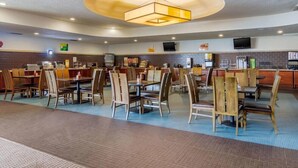 A spacious restaurant with multiple dining tables and chairs, a central light fixture, and a kitchen area in the background.