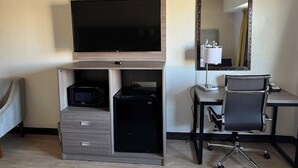 A hotel room with a flat-screen TV, a desk with a computer, a chair, a microwave, and a small refrigerator.