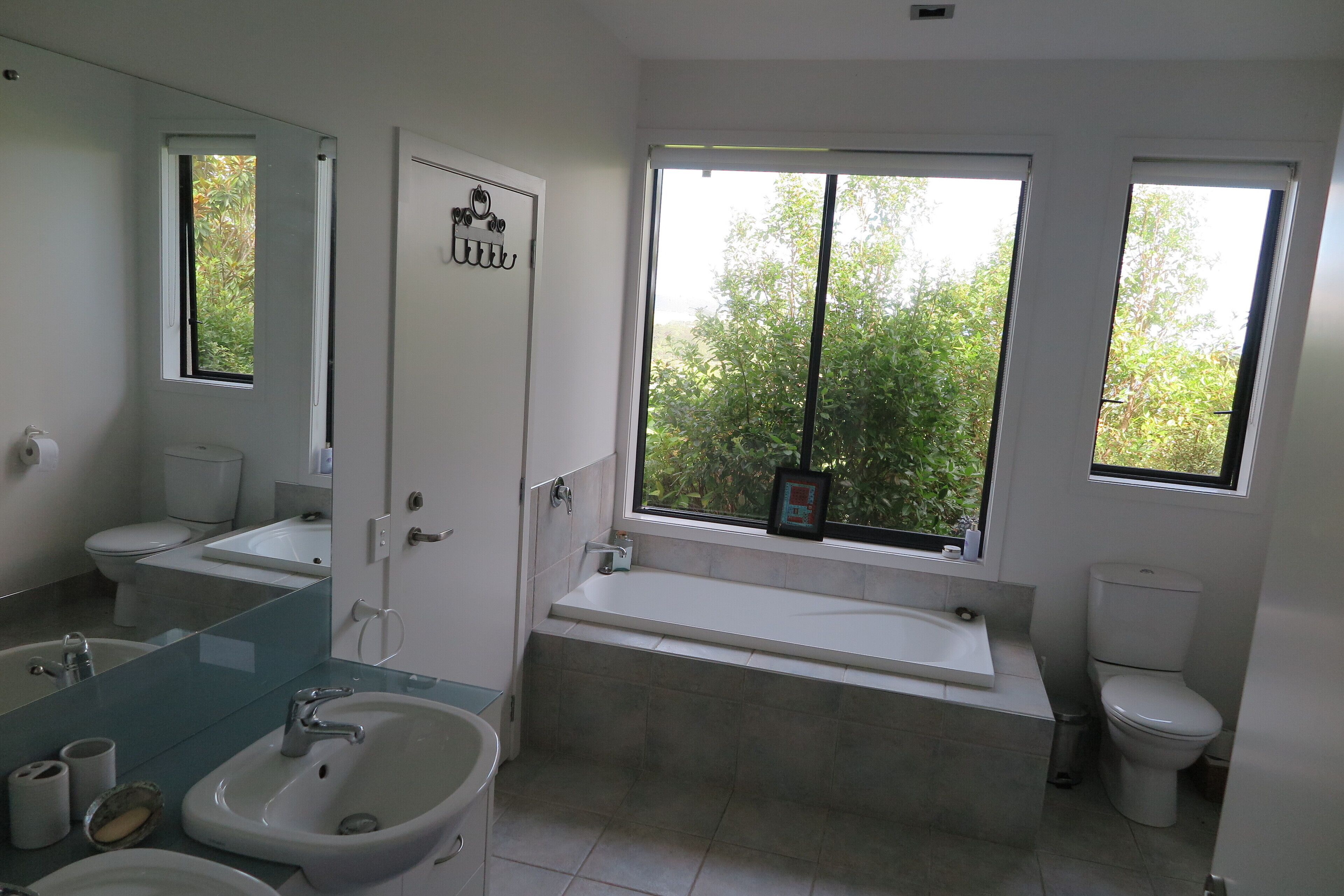 A modern bathroom with a bathtub, two sinks, a toilet, and a window providing a view of greenery.