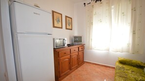 Une cuisine compacte équipée d’un réfrigérateur, d’un four à micro-ondes et d’un petit téléviseur posé sur un meuble en bois. Deux tableaux encadrés ornent le mur, et une fenêtre est agrémentée de rideaux.