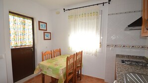 Un coin repas avec une table recouverte d’une nappe verte, des chaises en bois, une fenêtre agrémentée d’un rideau coloré et une cuisine équipée d’une cuisinière et d’un plan de travail carrelé.