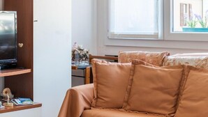 Ein gemütliches Zimmer mit einem beigefarbenen Sofa, einem Holzregal, einem Flachbildfernseher und einem Fenster mit Blick auf ein Gebäude.