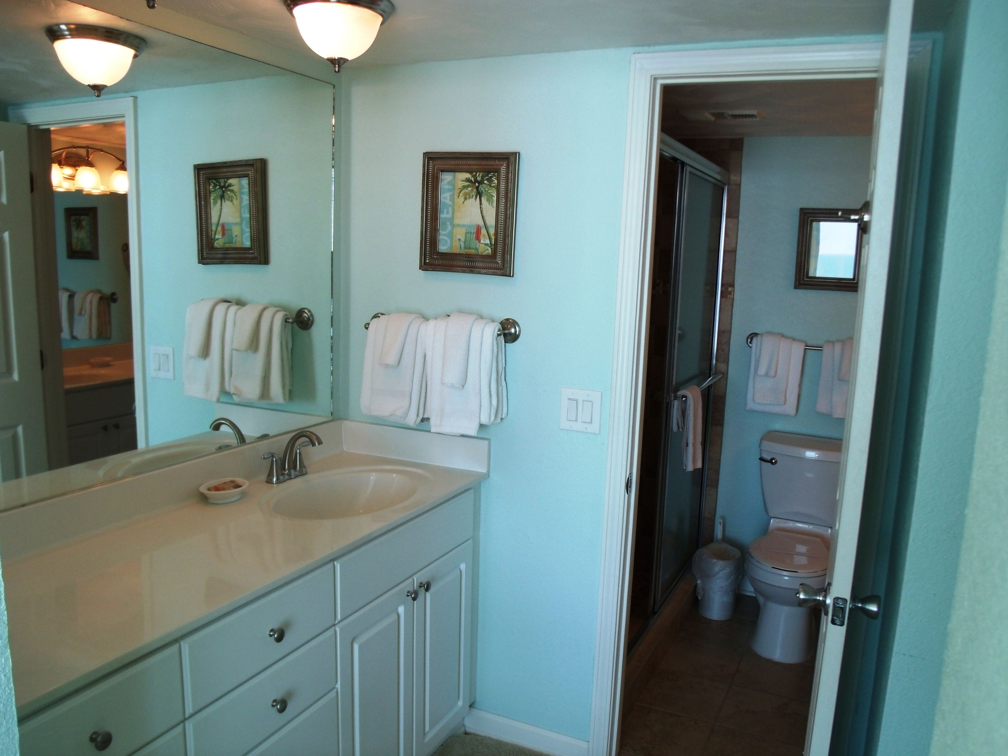 A bathroom with a double sink, a toilet, and a shower. There are towels hanging on the wall and a trash can.