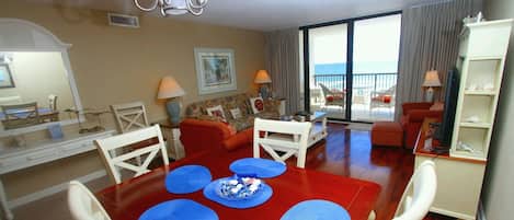 A dining area with a red table, white chairs, and blue placemats. There is a balcony with a view of the sea.