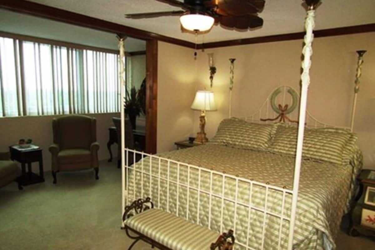 Una cama con dosel y cortinas, una mesita de noche con lámpara, una silla y una pequeña mesa con una planta.