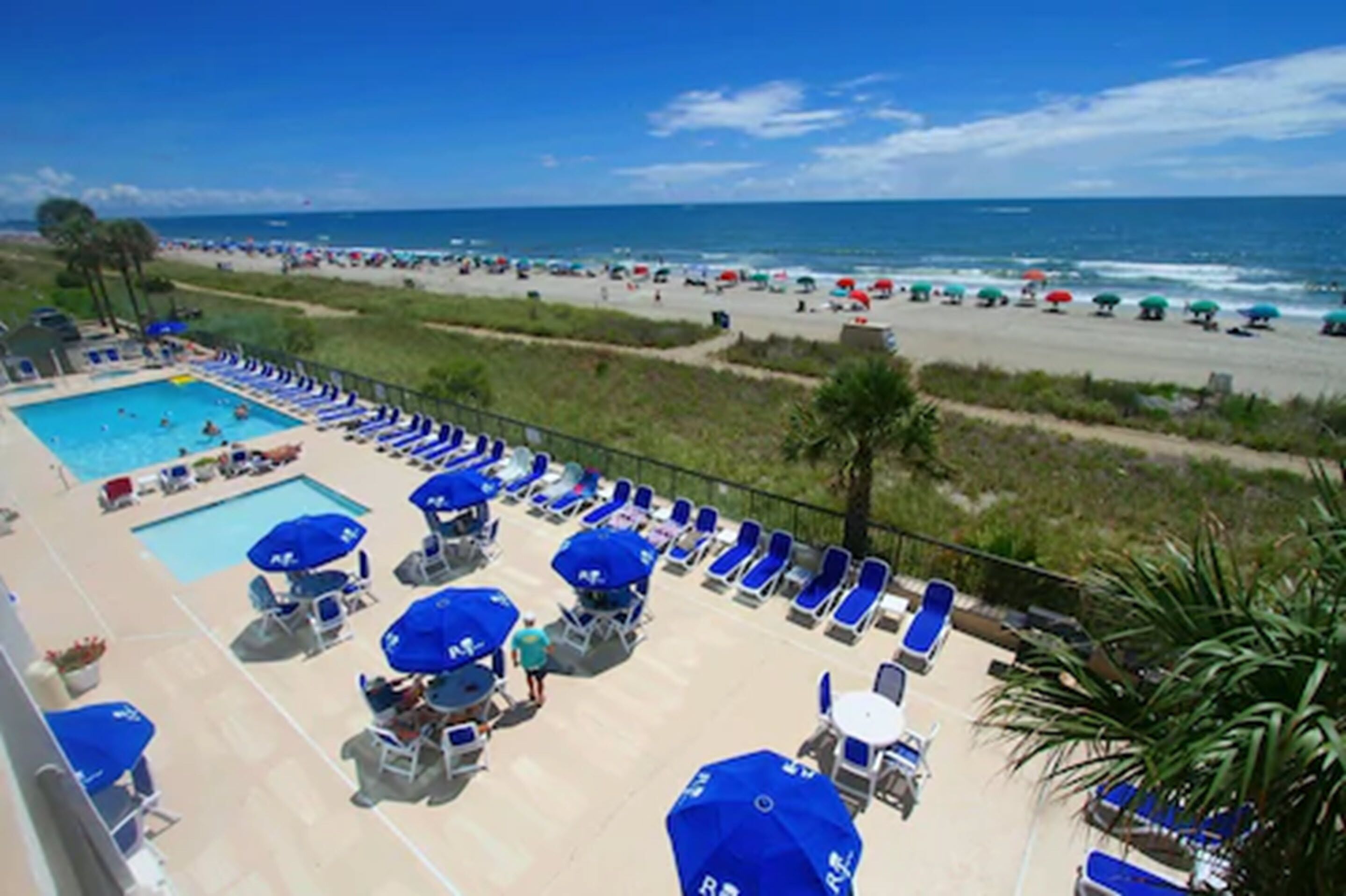 A pool area with blue umbrellas and lounge chairs, overlooking a beach with umbrellas and people.