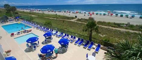 A pool area with blue umbrellas and lounge chairs, overlooking a beach with umbrellas and people.