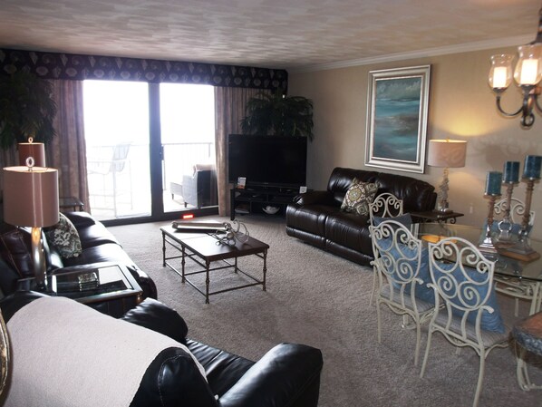 A living room with a sofa, coffee table, and TV. There is a window with curtains, a lamp, and a painting on the wall. - Regency Towers (Myrtle Beach)