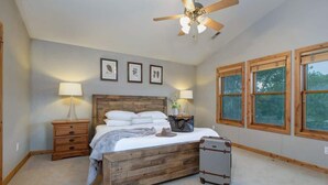 Une chambre avec un lit en bois, un ventilateur de plafond, des tableaux encadrés accrochés au mur et de grandes fenêtres.