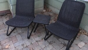 Två svarta fåtöljer av rotting med metallben, placerade på en stenlagd uteplats intill en byggnad med grön fasadbeklädnad.
