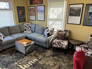 A living room with a grey sofa, a patterned ottoman, and a chair with a cow print cushion. There are framed pictures on the wall and a window with blinds.