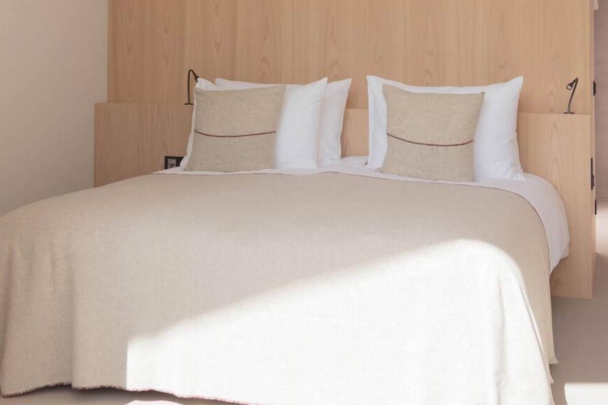 A bed with a beige bedspread and pillows, a wooden headboard, and a bedside table with a lamp.