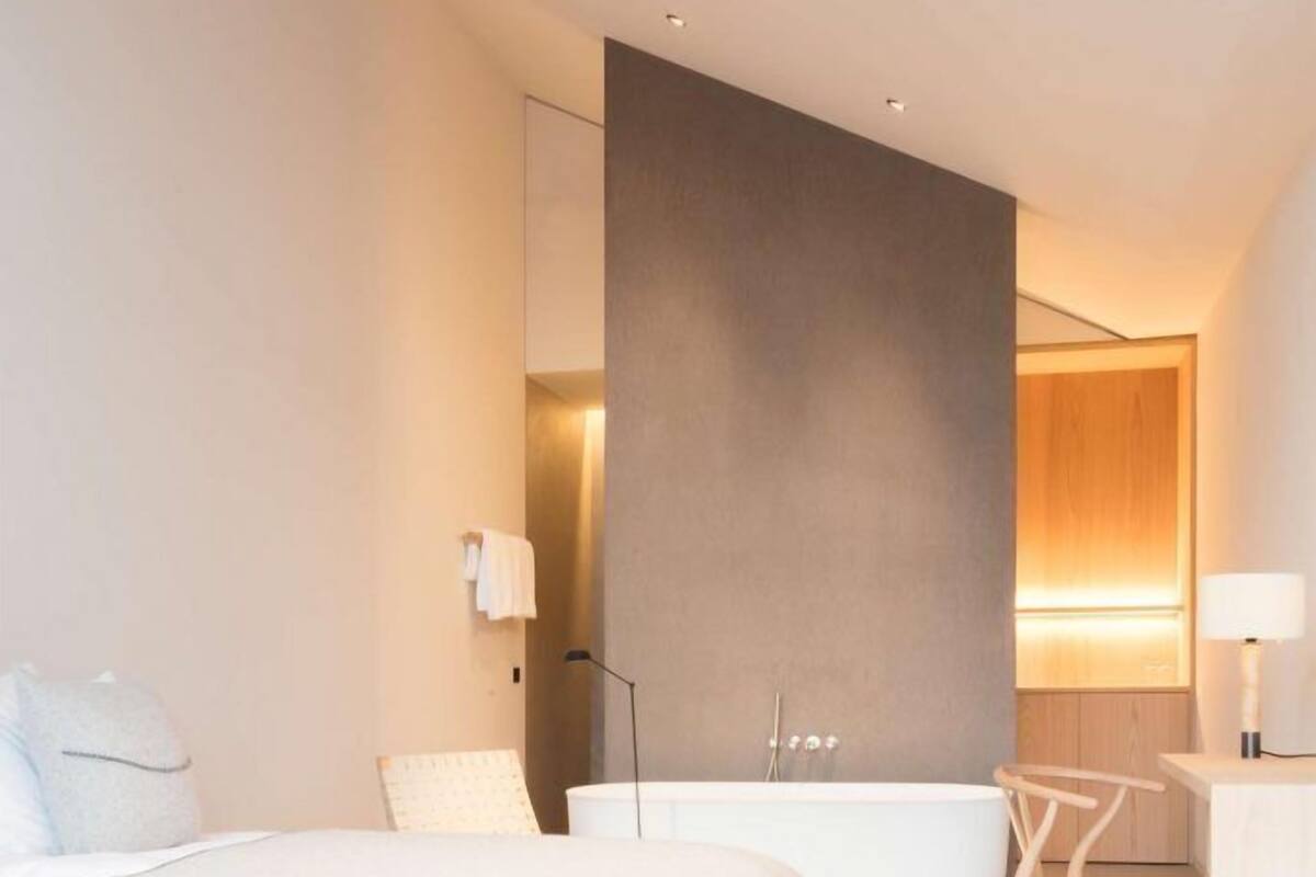 A modern hotel room with a large bed, a sitting area with a chair, a desk, and a bathtub.