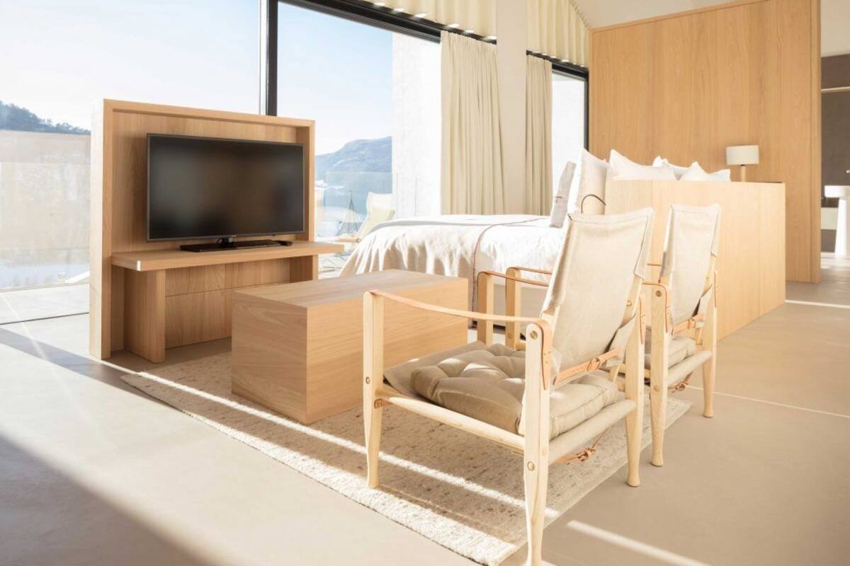A modern hotel room with a large flat-screen TV, a wooden desk, a comfortable chair, a bed with white linens, and a view of the mountains through large windows.