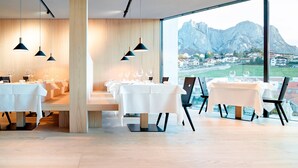 A modern dining area with tables set for a meal, large windows offering a view of a mountain and town, and pendant lighting. - Schgaguler Hotel (Castelrotto)
