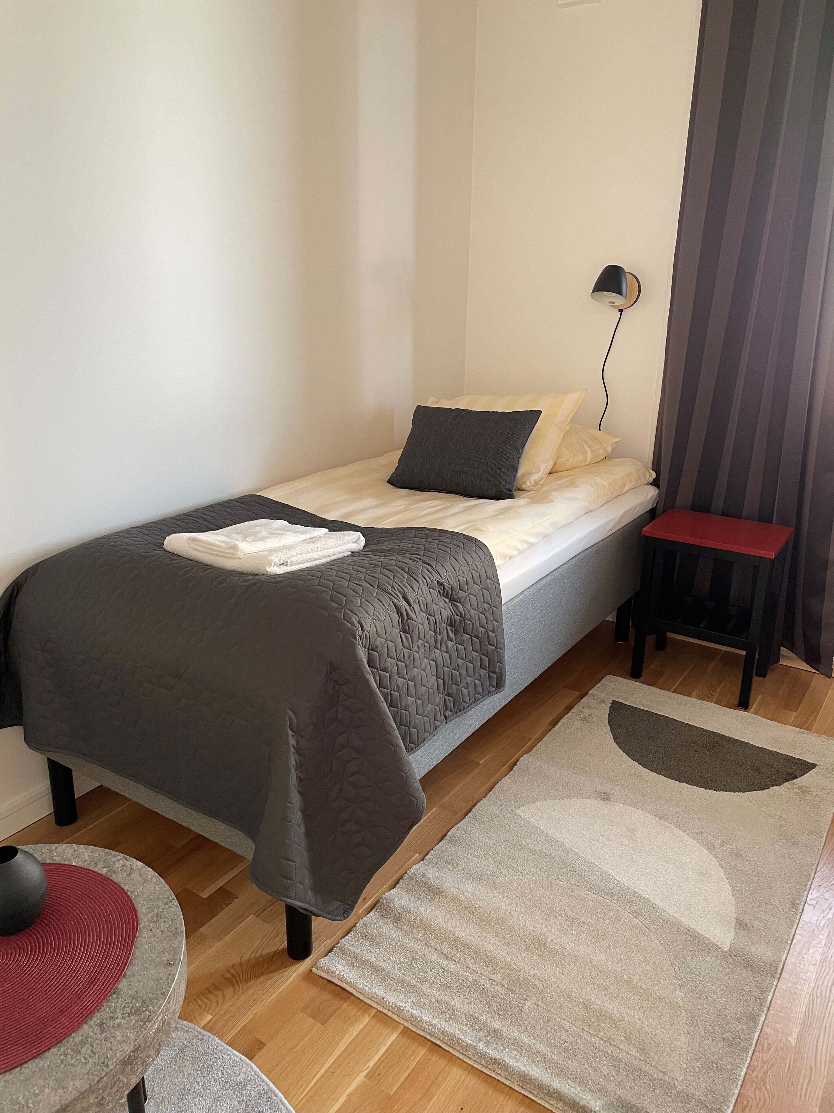 Un dormitorio pequeño y ordenado con una cama individual, una mesita de noche, una silla y una lámpara de pie.