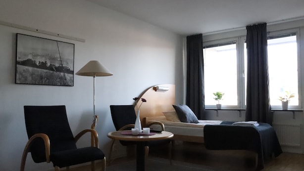 A hotel room with a bed, two chairs, a small table with a lamp, a painting on the wall, and a window with curtains. - Räddningsskolans lägenhetshotell (Skovde)