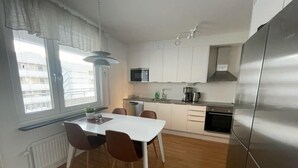 A compact kitchen with white cabinets, a stainless steel refrigerator, a dining table with chairs, and a window with blinds. - Räddningsskolans lägenhetshotell (Skovde)