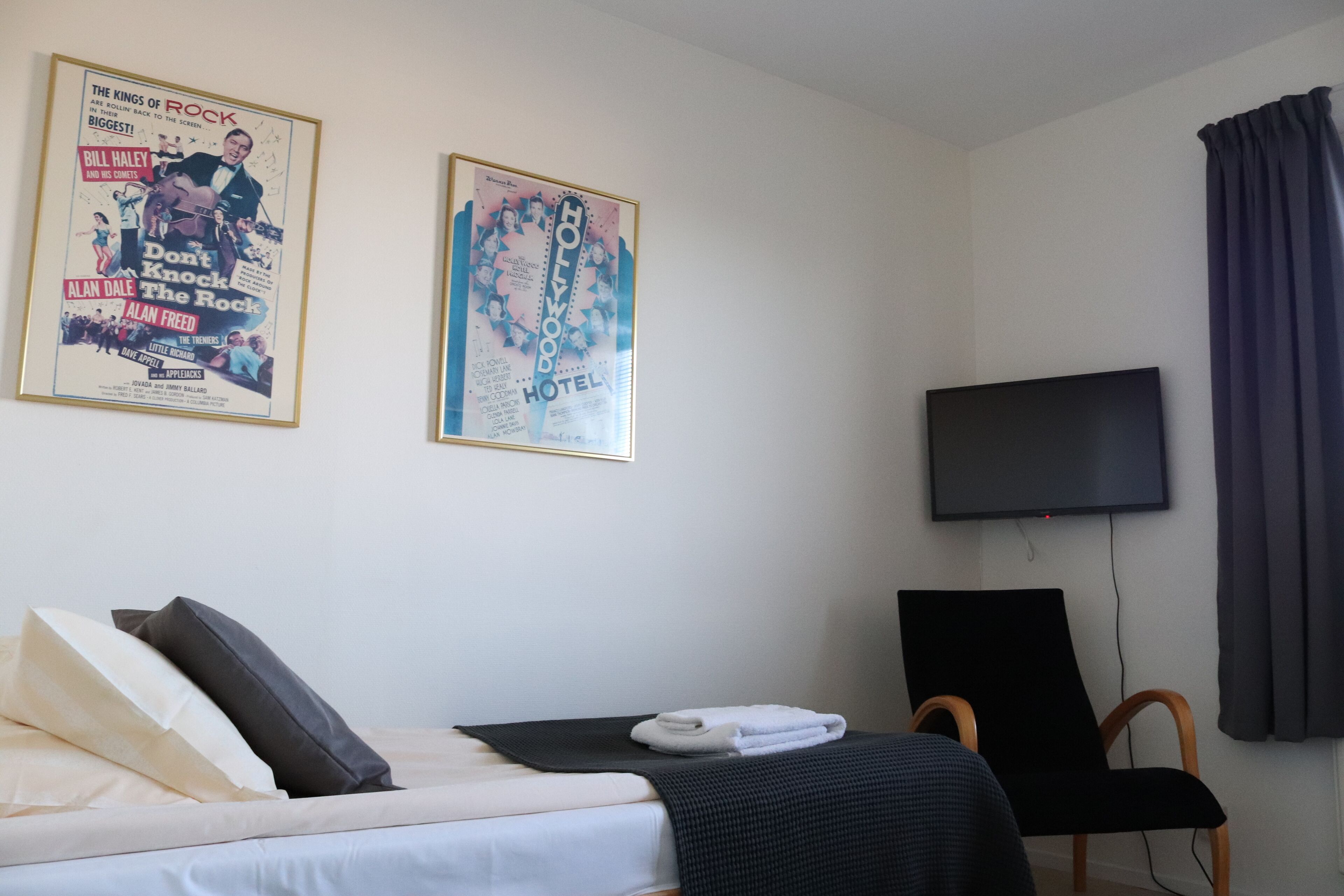 A hotel room with a bed, a flat-screen TV, a black chair, and two posters on the wall.