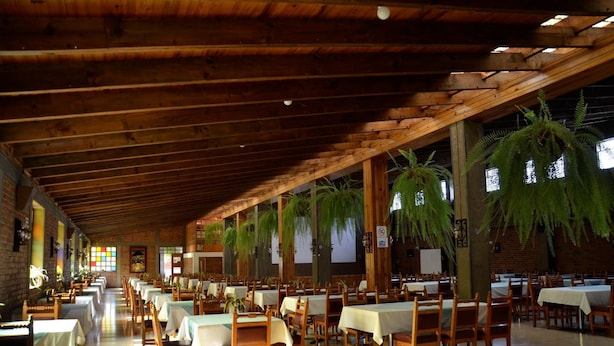 A spacious indoor dining area with wooden beams, tables set with white tablecloths, and hanging ferns. - Hotel del Campo (Quetzaltenango)