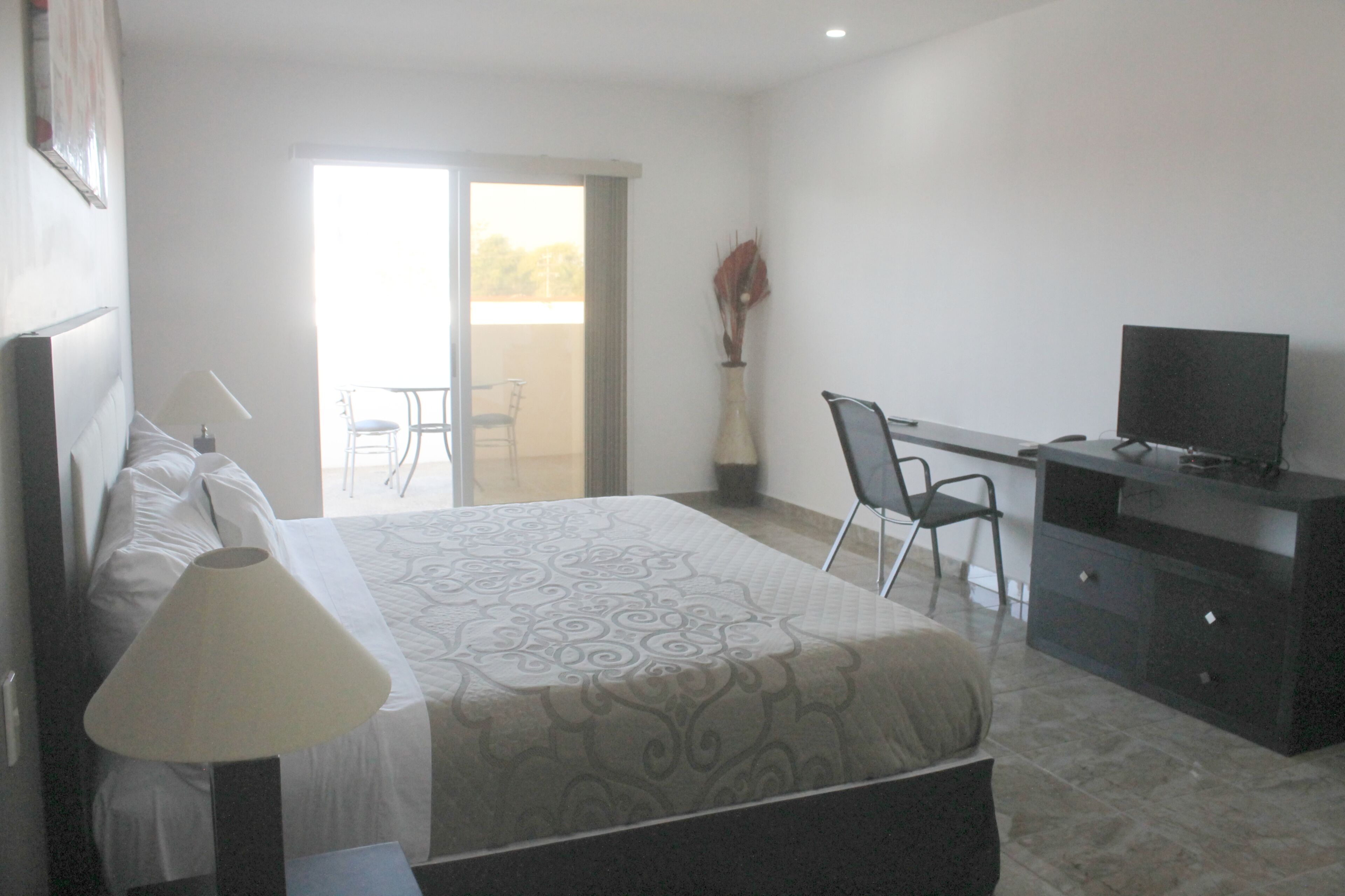A hotel room with a bed, a desk with a TV, a chair, and a balcony with a chair.