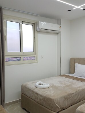 A bedroom with a bed, a window, an air conditioner, and a view of the cityscape.