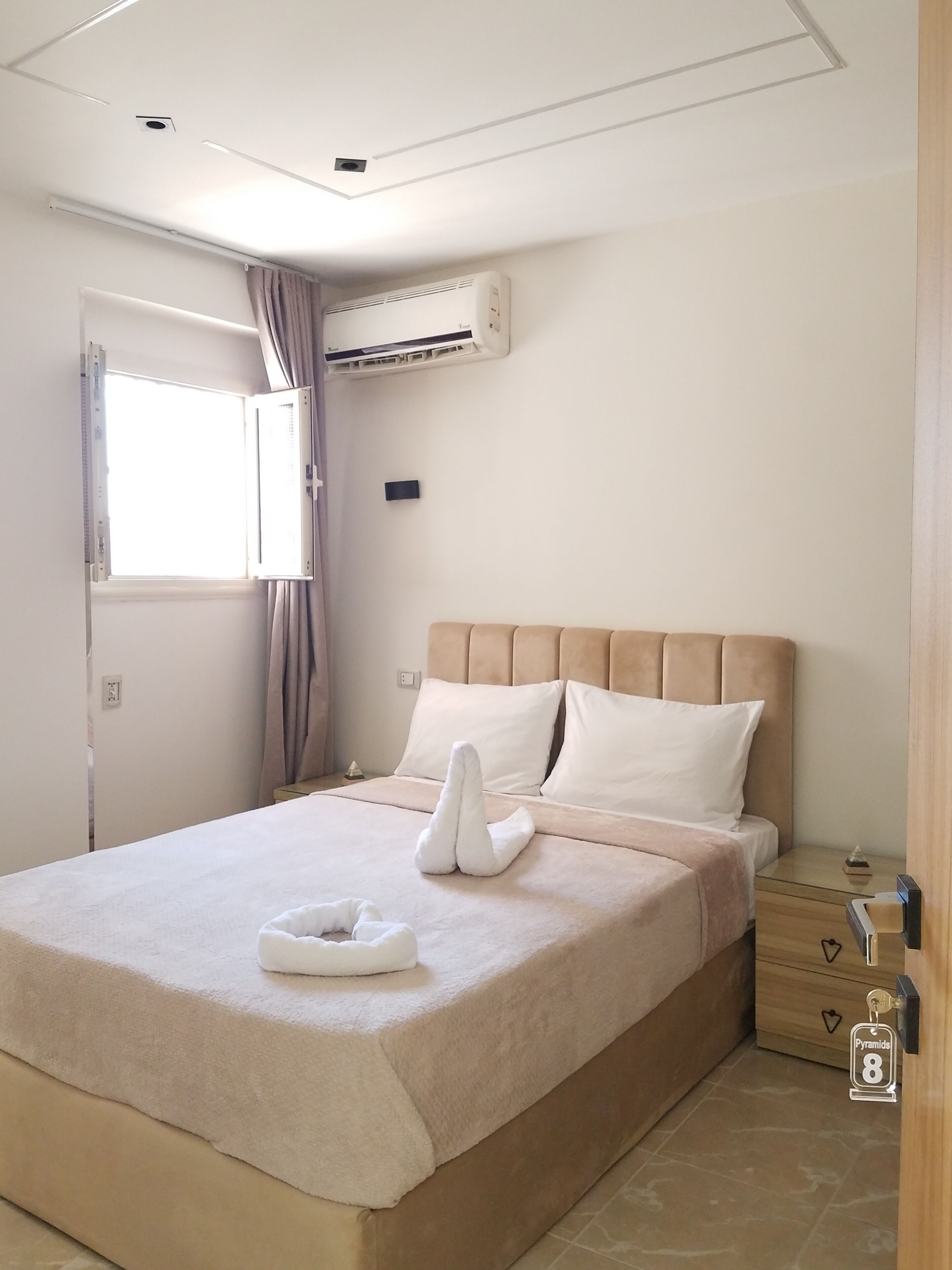 A hotel room with a bed, a window, an air conditioning unit, a bedside table, and a nightstand.