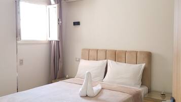 A hotel room with a bed, a window, an air conditioning unit, a bedside table, and a nightstand.