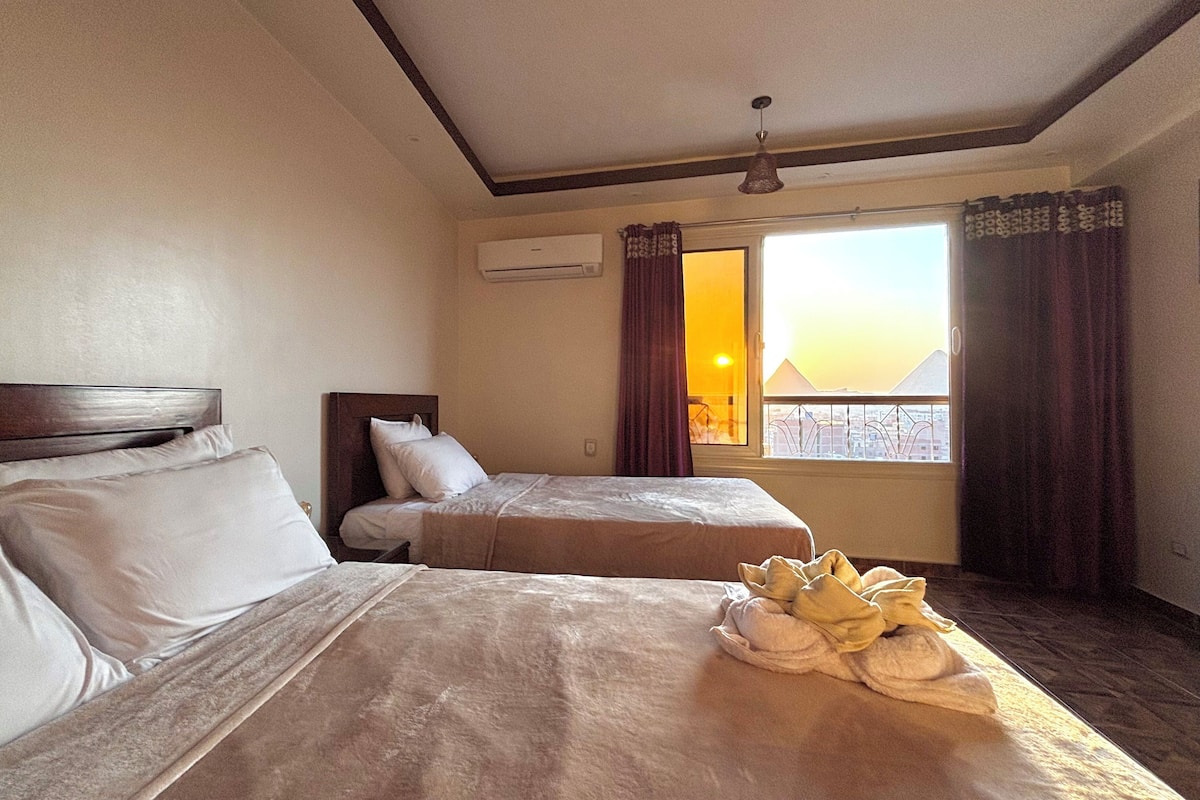 A hotel room with two beds, a window with curtains, an air conditioning unit, and a view of a cityscape.