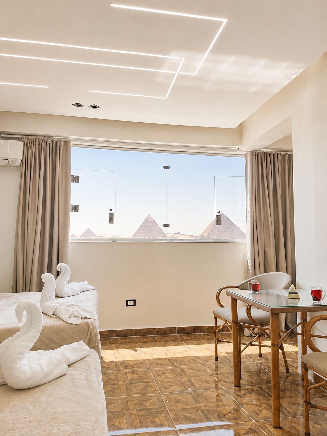 A hotel room with a bed, a table with chairs, a view of pyramids, and a modern ceiling design.