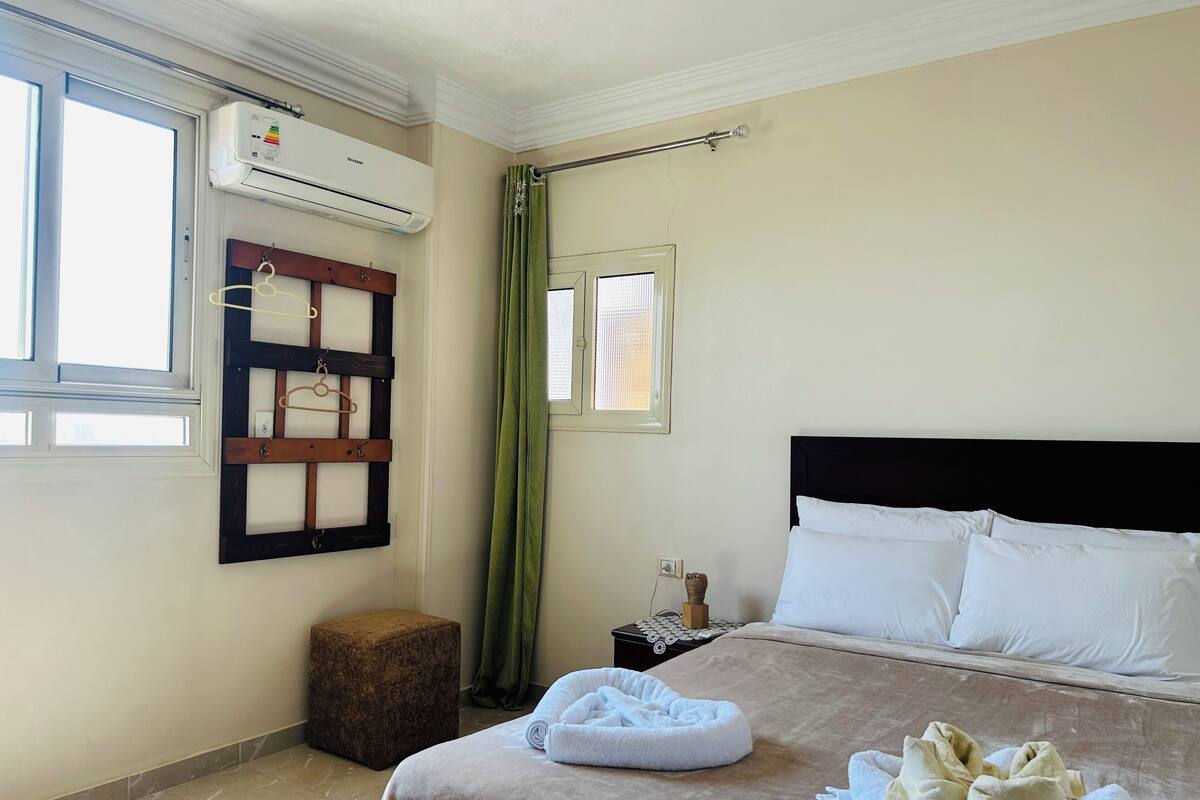 A hotel room with a bed, a small bedside table, a green curtain, and an air conditioning unit.