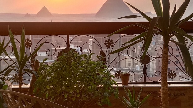 A balcony with a view of the pyramids at sunset, potted plants, and a decorative chair. - Pyramids Terrace INN (Giza)