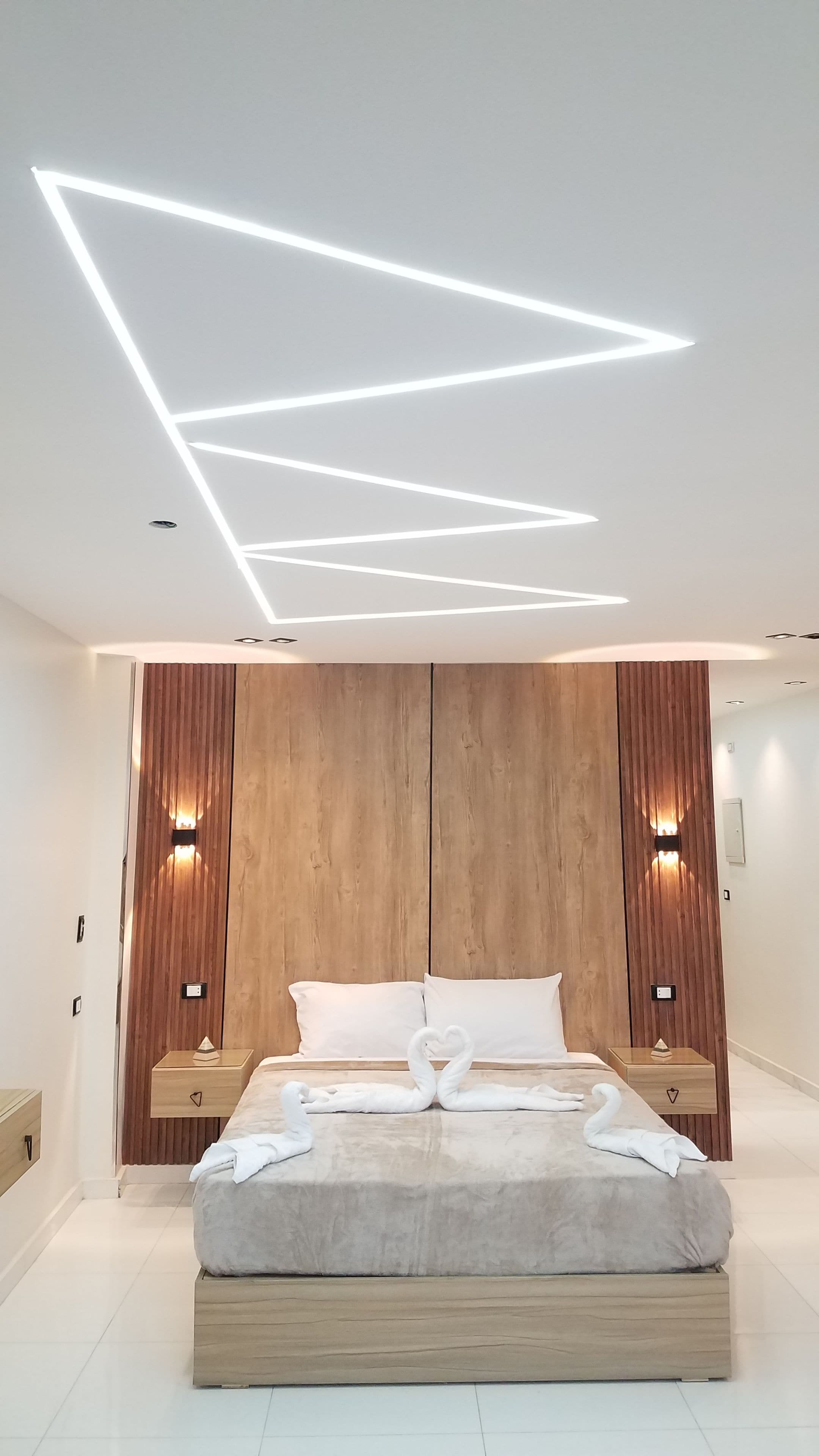 A modern bedroom with a geometric ceiling light design, a wooden panel wall, a bed with a heart-shaped towel arrangement, and bedside tables with wall-mounted lamps.