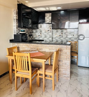A kitchen with a dining area, featuring a marble-tiled floor, wooden chairs, a table, black cabinets, and a white refrigerator. - Pyramids Terrace INN (Giza)