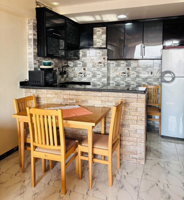 A kitchen with a dining area, featuring a marble-tiled floor, wooden chairs, a table, black cabinets, and a white refrigerator. - Pyramids Terrace INN (Giza)