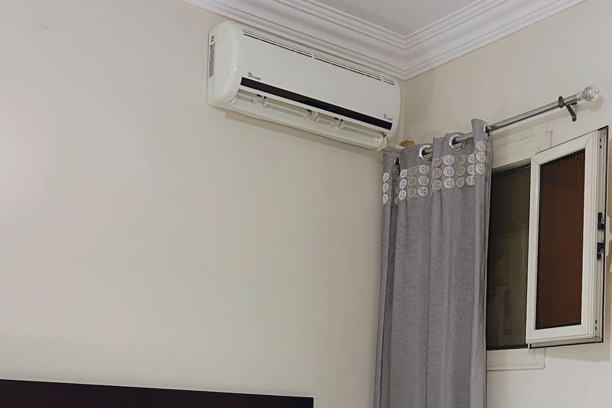 A hotel room with a bed, white pillows, a grey curtain, and an air conditioning unit.