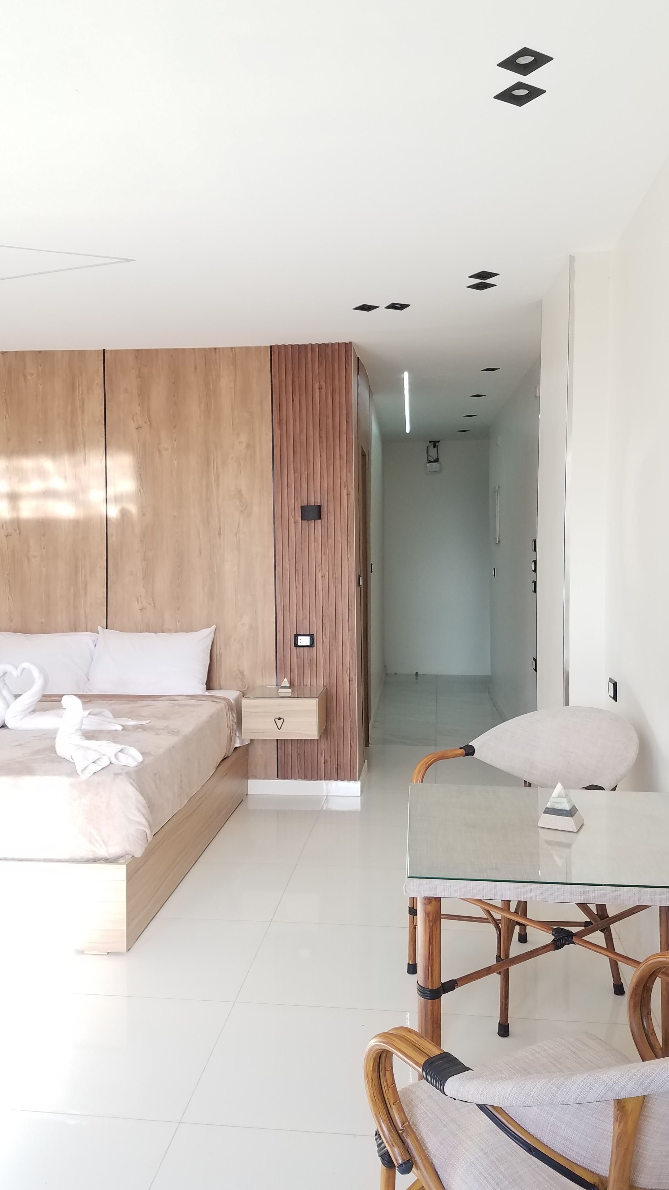 A modern bedroom with a wooden wardrobe, a bed with white linens, a bedside table, and a glass-top dining table with a chair.