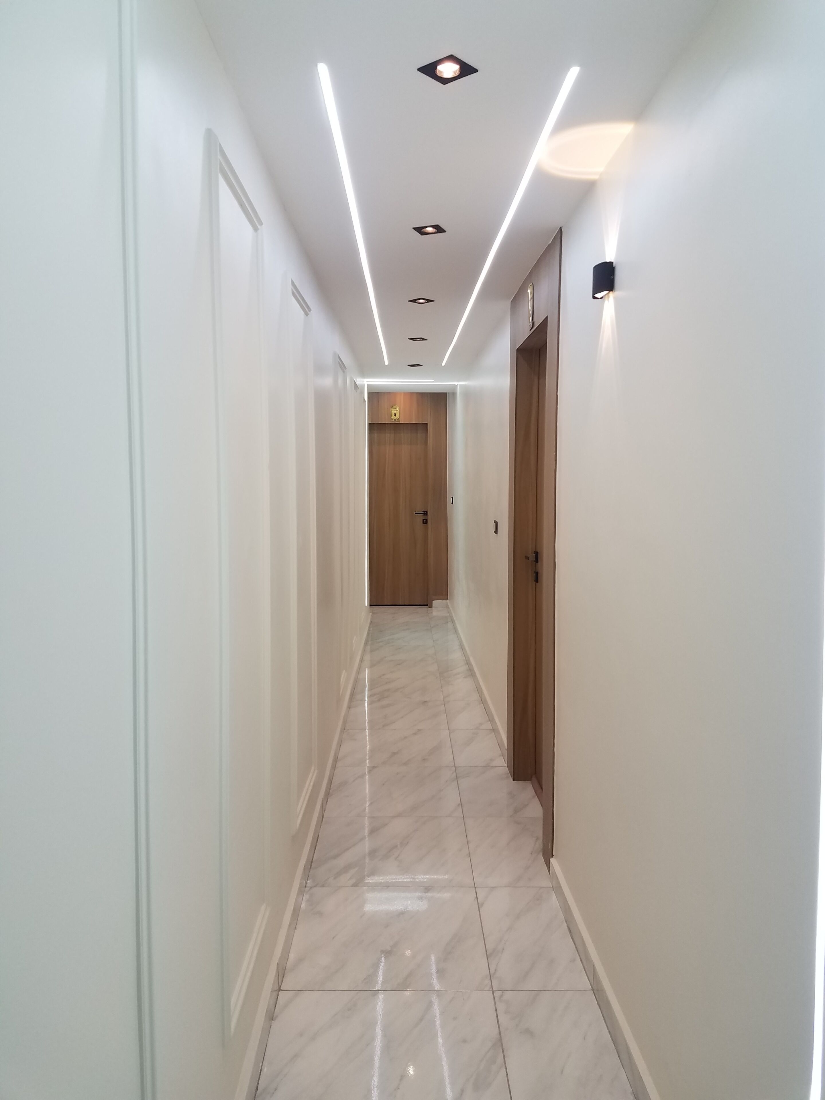 A corridor with white walls, a wooden door at the end, and a tiled floor.