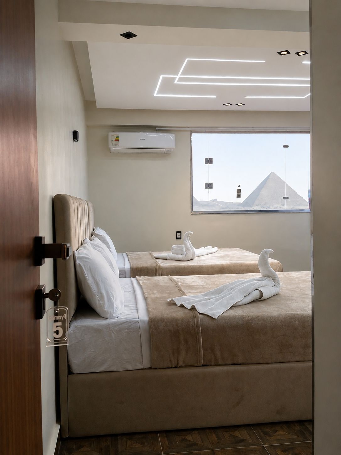 Habitación de hotel con dos camas, una ventana con vista a una pirámide, un equipo de aire acondicionado e iluminación de techo moderna.