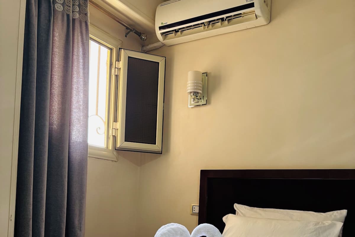 A hotel room with a bed, a window with curtains, an air conditioner, a wall-mounted light, and a heart-shaped towel arrangement.