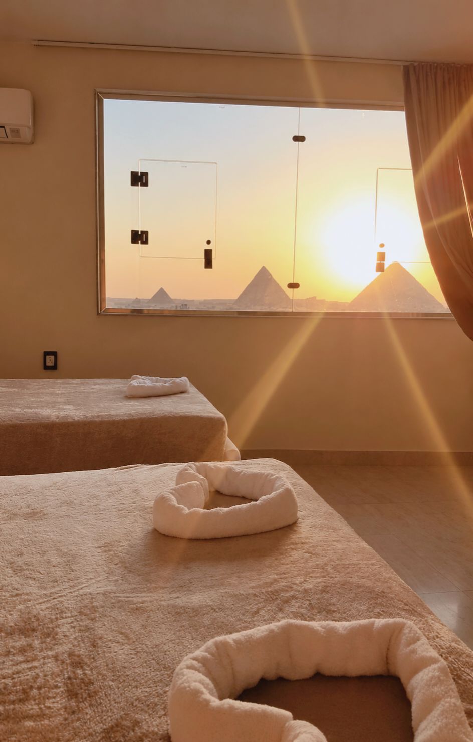 A room with two beds, a large window with a view of pyramids, and a sun setting.